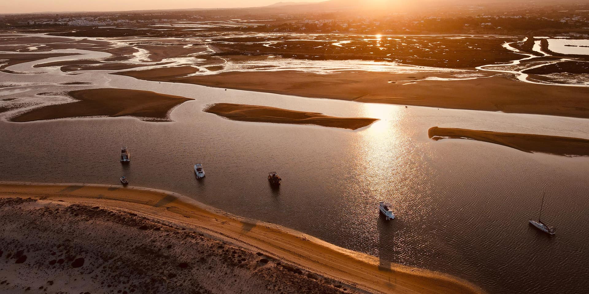 Vista aérea de uma ria ao pôr do sol, com barcos fundeados num canal entre bancos de areia.