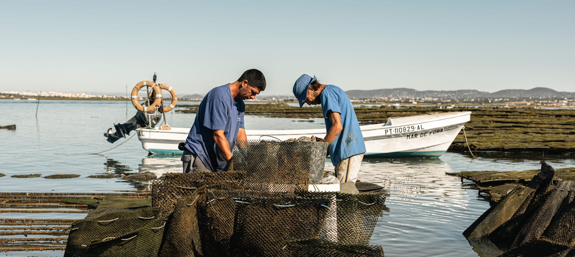 Dois mariscadores a trabalhar sobre cestos na Ria Formosa, com os barcos atracados atrás de si.