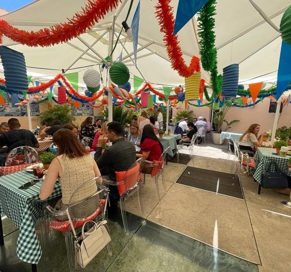 Um terraço de hotel decorado para os Santos Populares: balões de papel, grinaldas e bandeirinhas sobre mesas com toalhas aos quadrados e vasos de manjerico, com hóspedes à mesa.