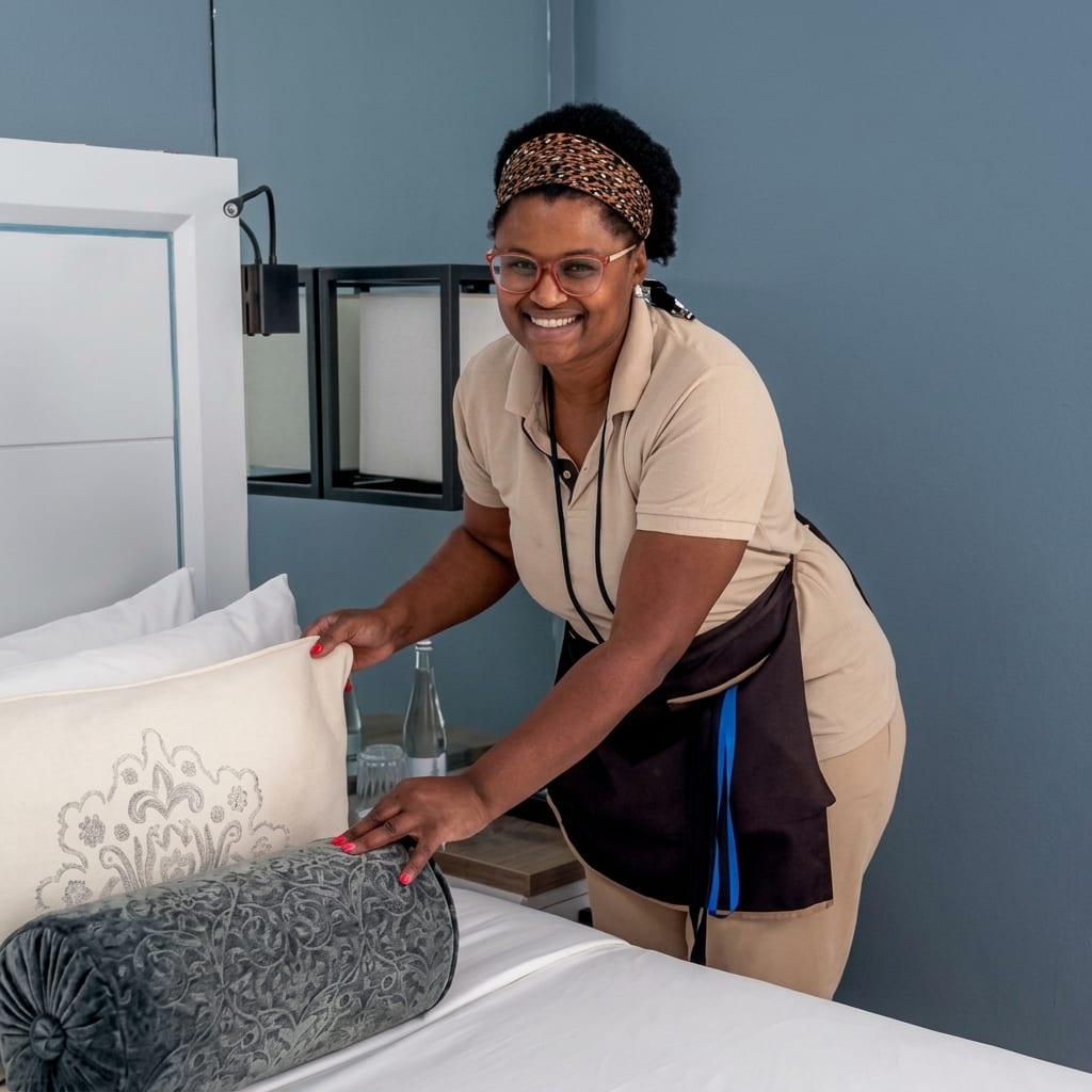 Uma colaboradora de andares, com o uniforme do Grupo, sorri enquanto coloca as almofadas numa cama feita.