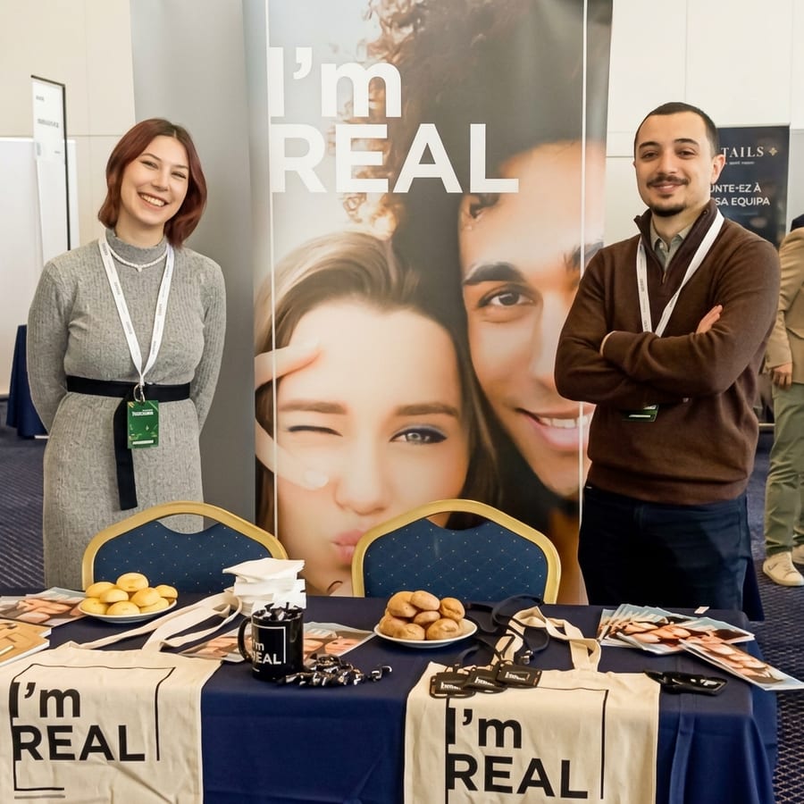 Dois colaboradores no stand de recrutamento “I’m REAL” do Grupo, junto a um cartaz e a uma mesa com materiais de marca.