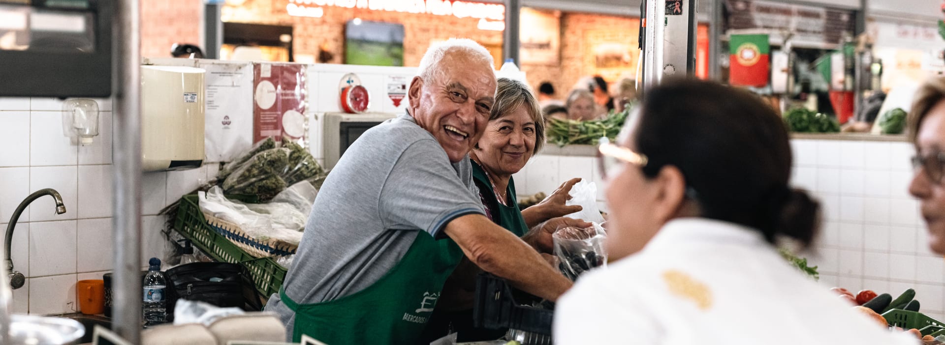 Comerciantes e uma chefe de hotel a cumprimentarem-se ao balcão de um mercado, com legumes e fruta empilhados.
