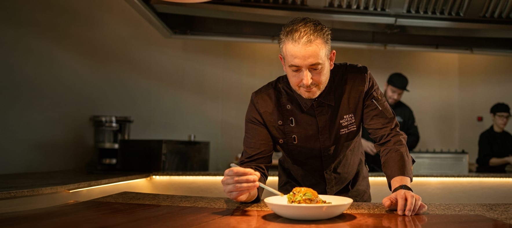 Um chefe do Grupo a empratar no passe de uma cozinha de hotel, com colegas a trabalhar atrás de si.