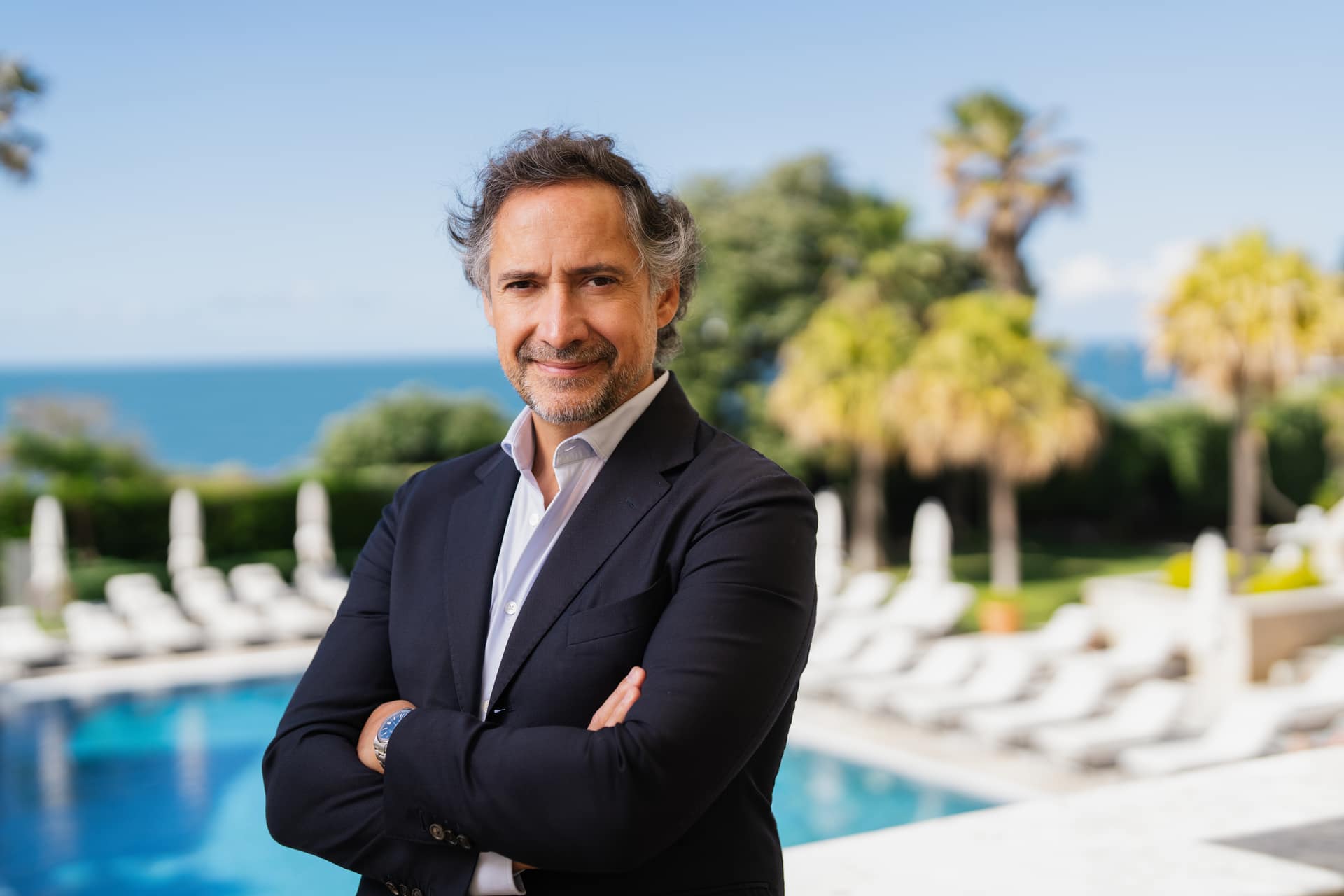 Eurico de Almeida, de braços cruzados, junto a uma piscina com o mar ao fundo.