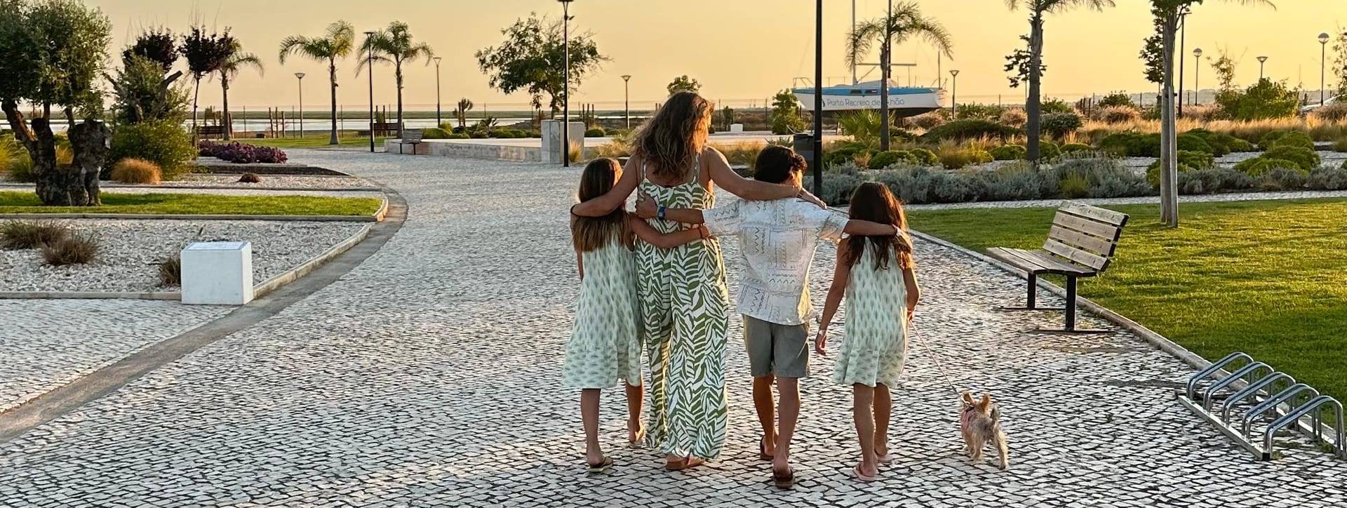 Uma família a caminhar de costas por um passeio marítimo em calçada, ao pôr do sol, com um cão pequeno ao lado.