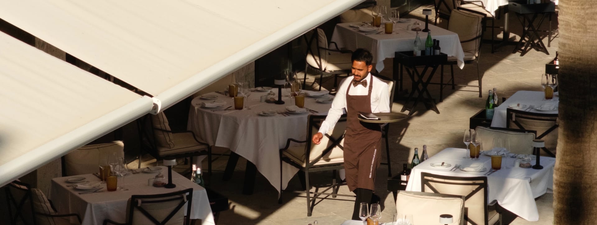 Um empregado de mesa a transportar um tabuleiro entre mesas postas, no terraço sombreado do restaurante de um hotel.