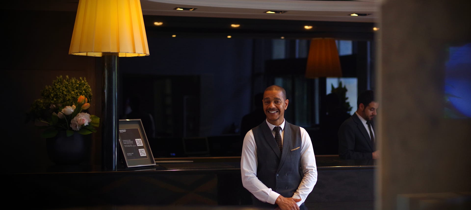Um concierge sorri atrás do balcão de receção de um hotel ao anoitecer, com candeeiros acesos ao longo do balcão.