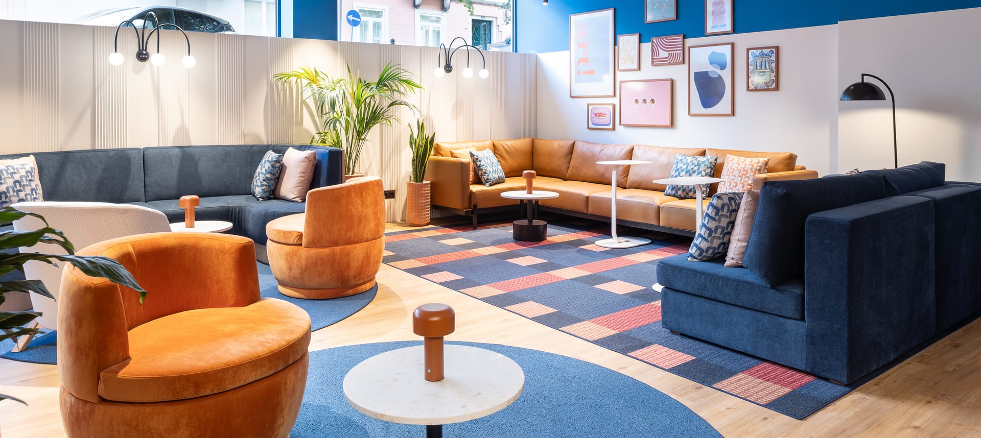 O lounge de um hotel do Grupo: sofás baixos em tons de caramelo e azul-marinho, um tapete padronizado e uma parede de gravuras emolduradas.