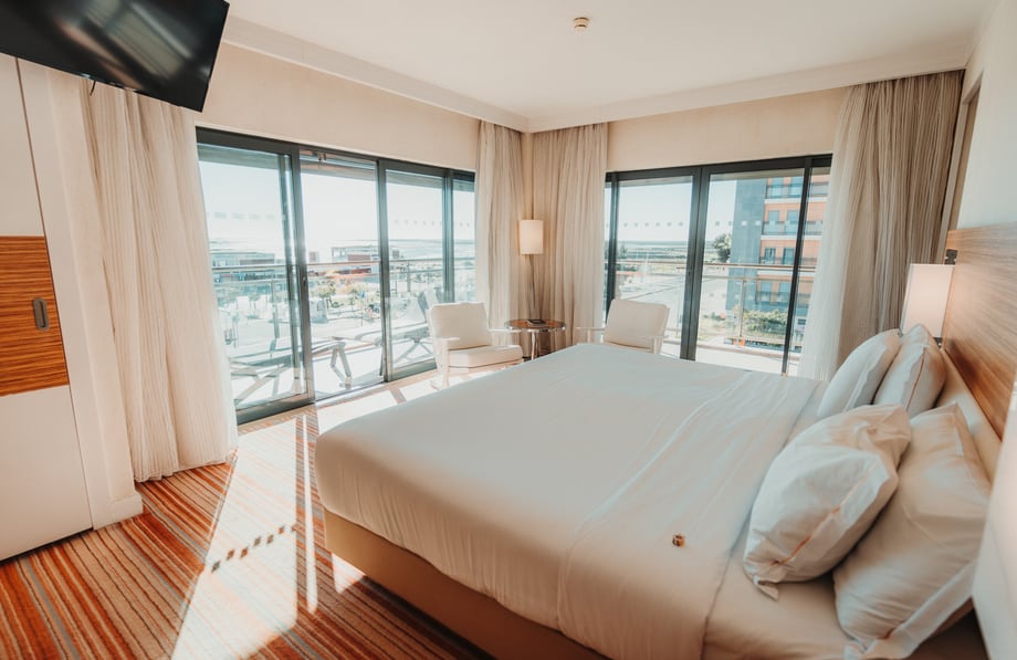 Um quarto de hotel com as cortinas abertas sobre uma varanda e o mar ao fundo.