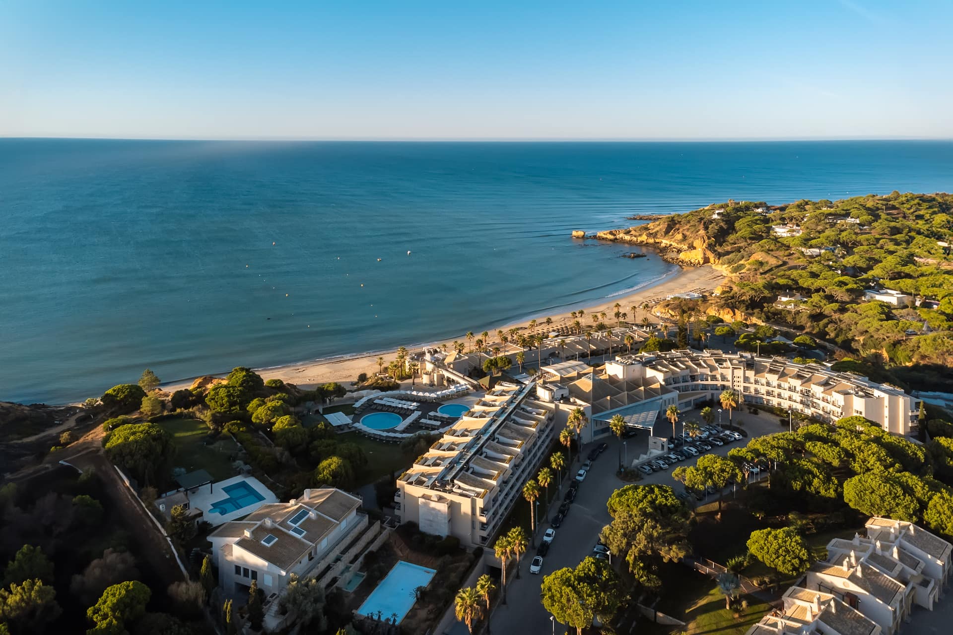 Vista aérea de um resort sobre a praia: piscinas entre os pinheiros e o mar ao fundo.