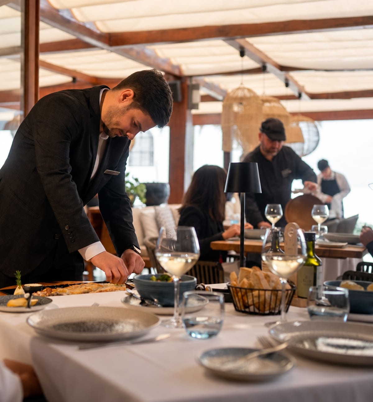 Um empregado a empratar peixe à mesa num restaurante do Grupo, com a equipa de cozinha a trabalhar ao fundo.